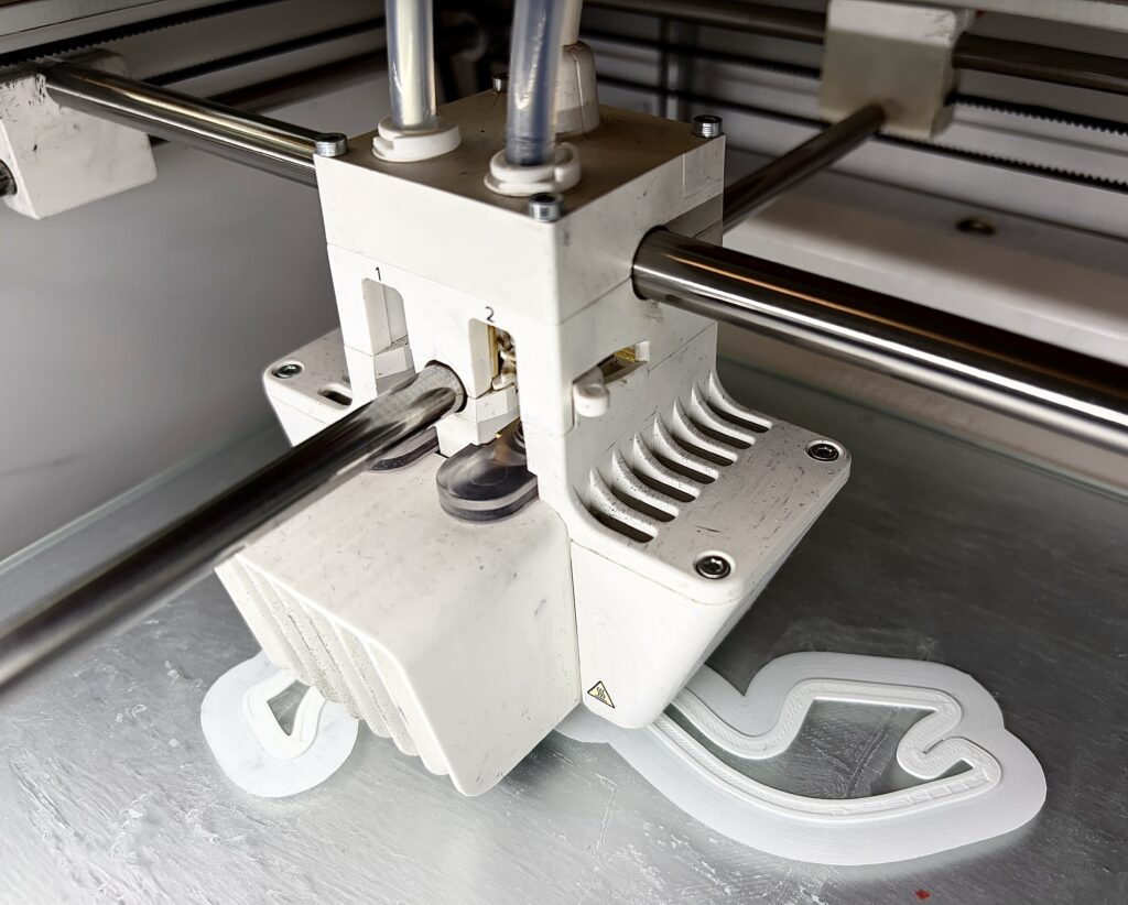 The print head of one of the Makerspace 3D printers depositing white PLA filament layer by layer onto the build plate. Students can bring their own designs or learn to model from scratch with help from a trained student ambassador. (Singh, 2026)